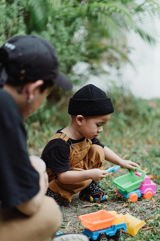Toddler in a beanie and overalls playing with brightly colored plastic trucks outside