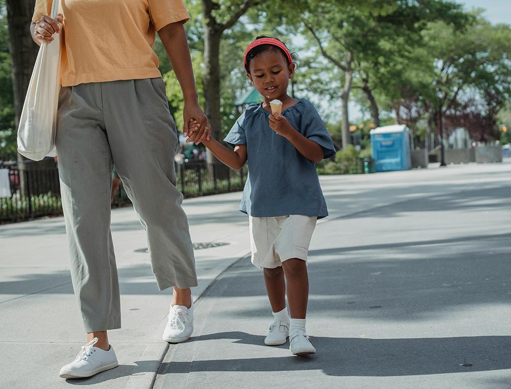 Young girl eating an ice cream cone while walking on a city street with her mother